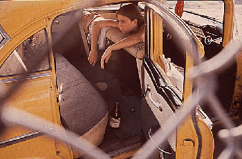Photograph of a teenage girl sitting backwards in a car with the doors open with her arms over the seat as seen through a chain link fence.